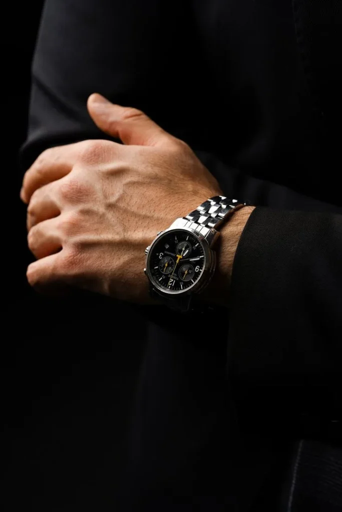 A close-up of a man wearing a Tissot watch with a black chronograph dial and stainless steel bracelet, styled with a formal black suit.