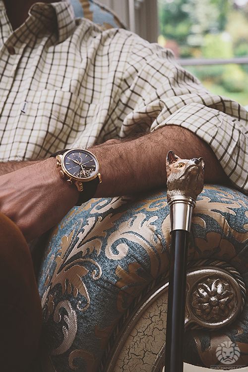 A man wearing a checkered button-down shirt and a luxury gold tourbillon chronograph analog watch, sitting on a blue damask upholstered chair next to a decorative walking cane with a fox head handle.