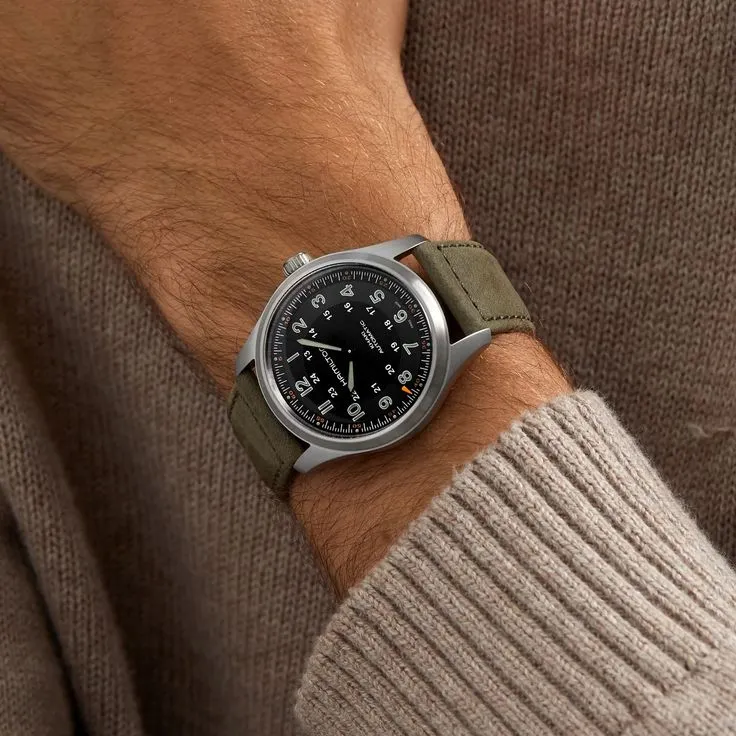 A man wearing a Hamilton Khaki Field automatic wristwatch with a black dial and olive green textile strap, styled with a brown knit sweater to showcase a versatile timepiece for any watch collection.