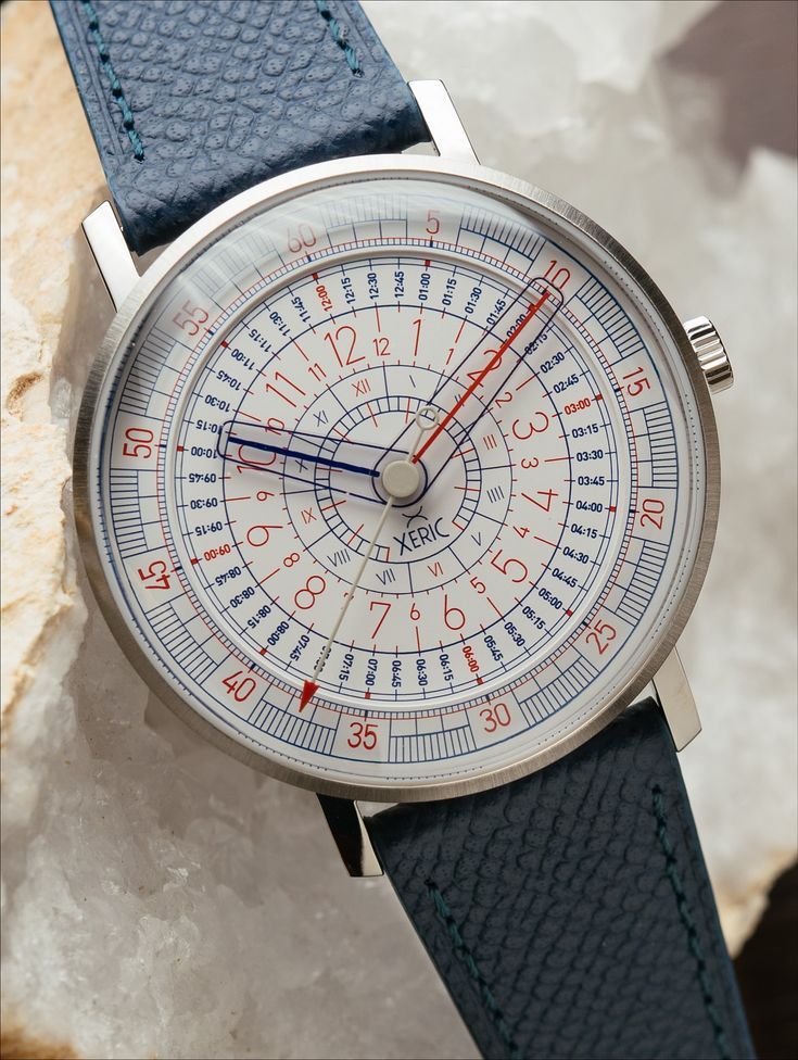 Xeric Omnigraph watch resting on white mineral rock, featuring an intricate multi-ring dial with concentric red and blue time tracks and sapphire crystal hands.