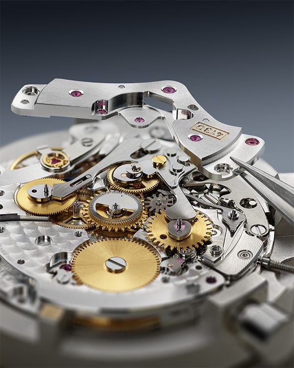 Watchmaker assembling a Rolex Calibre 4130 mechanical chronograph movement with tweezers, displaying brass gears, bridges, and ruby jewels.