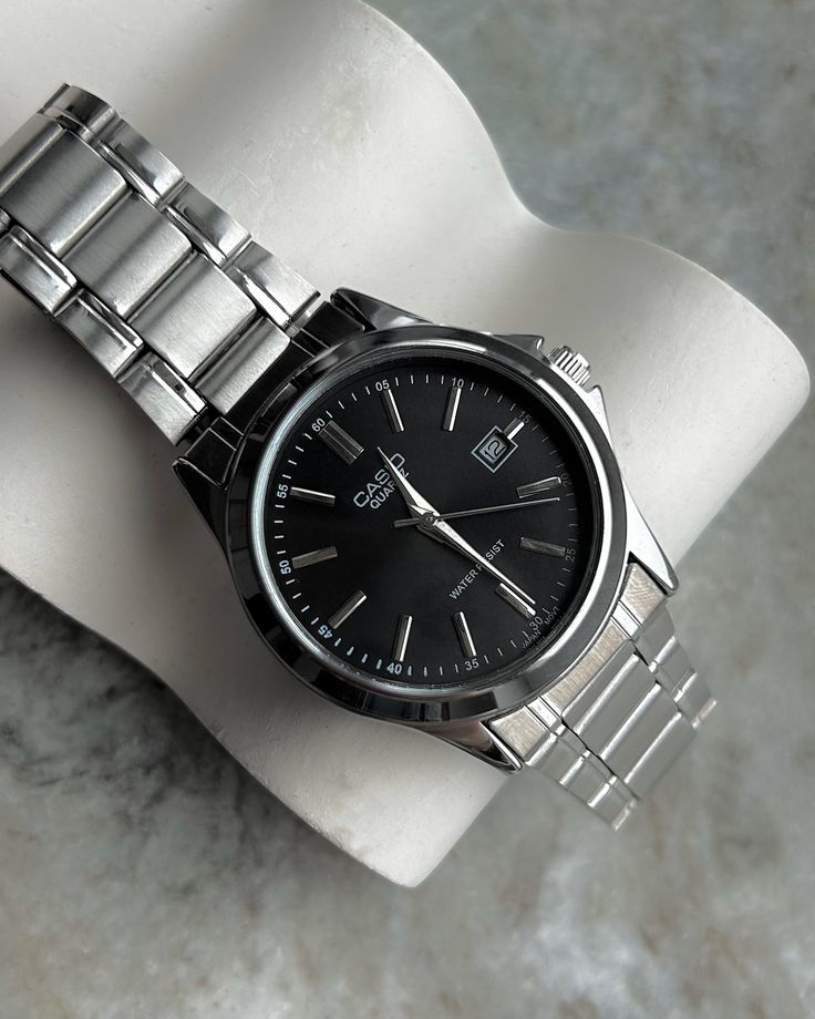 Casio Quartz dress-sports watch featuring a black dial, silver baton indices, date display window, and a stainless steel link bracelet resting on a white display holder.